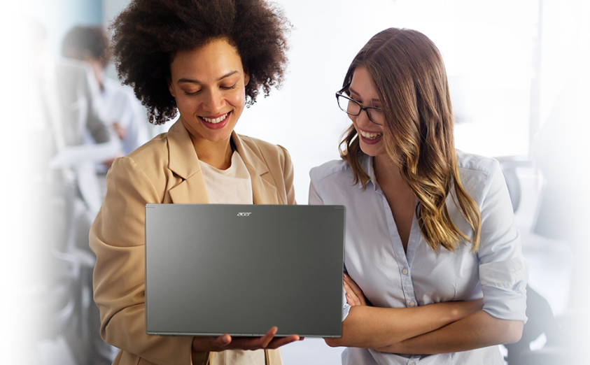 Uma das mulheres segura um notebook Acer enquanto a outra olha para a tela, ambas aparentam estar discutindo algo  relacionado ao conteúdo na tela do computador.