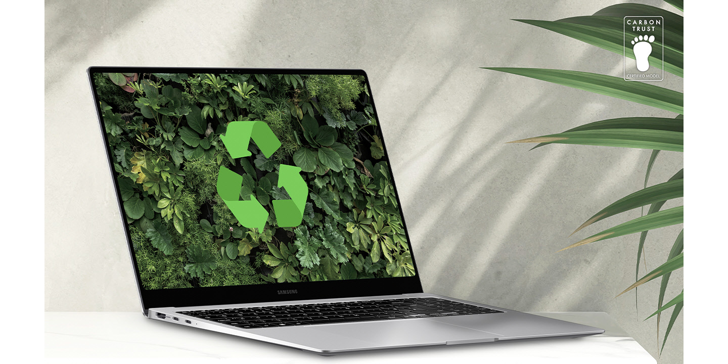 Galaxy Book4 Pro cinza posicionado sobre uma mesa ao lado de uma planta verde, aberto e voltado levemente para a direita com um papel de parede verde exuberante e logotipo de reciclagem mostrado na tela. Logotipo da certificação da Carbon Trust com o texto “CARBON TRUST. MODELO CERTIFICADO.