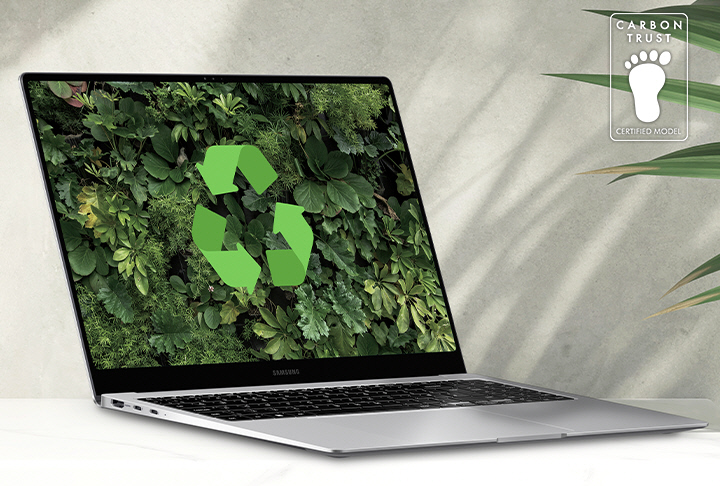 Galaxy Book4 Pro cinza posicionado sobre uma mesa ao lado de uma planta verde, aberto e voltado levemente para a direita com um papel de parede verde exuberante e logotipo de reciclagem mostrado na tela. Logotipo da certificação da Carbon Trust com o texto “CARBON TRUST. MODELO CERTIFICADO.”