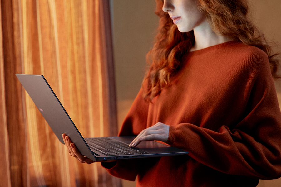 Uma jovem em pé em um ambiente fechado segurando o Galaxy Book4 Cinza aberto enquanto digita no teclado do laptop.