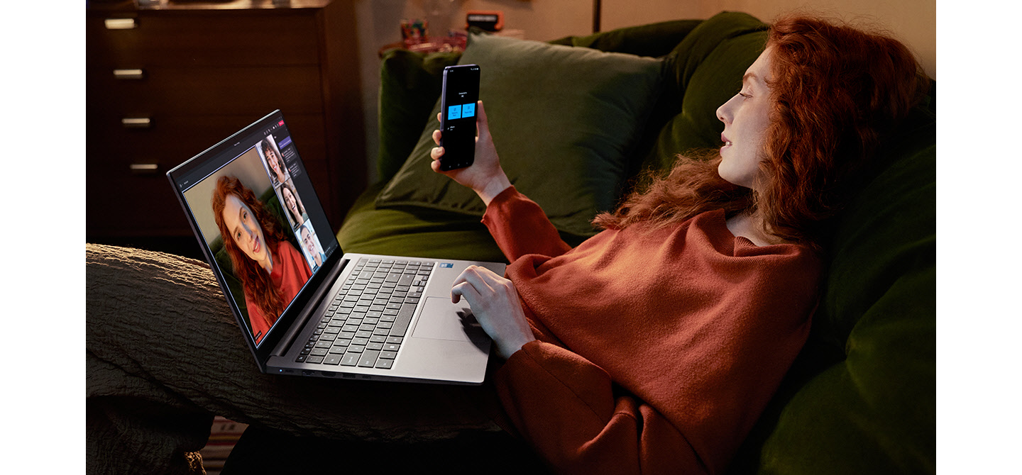 Uma jovem sentada em um sofá em casa está usando o Galaxy S24 Plus como uma câmera conectada com o Galaxy Book4 Cinza para participar de uma chamada em vídeo em grupo do Microsoft Teams com três outras pessoas mostradas na tela.